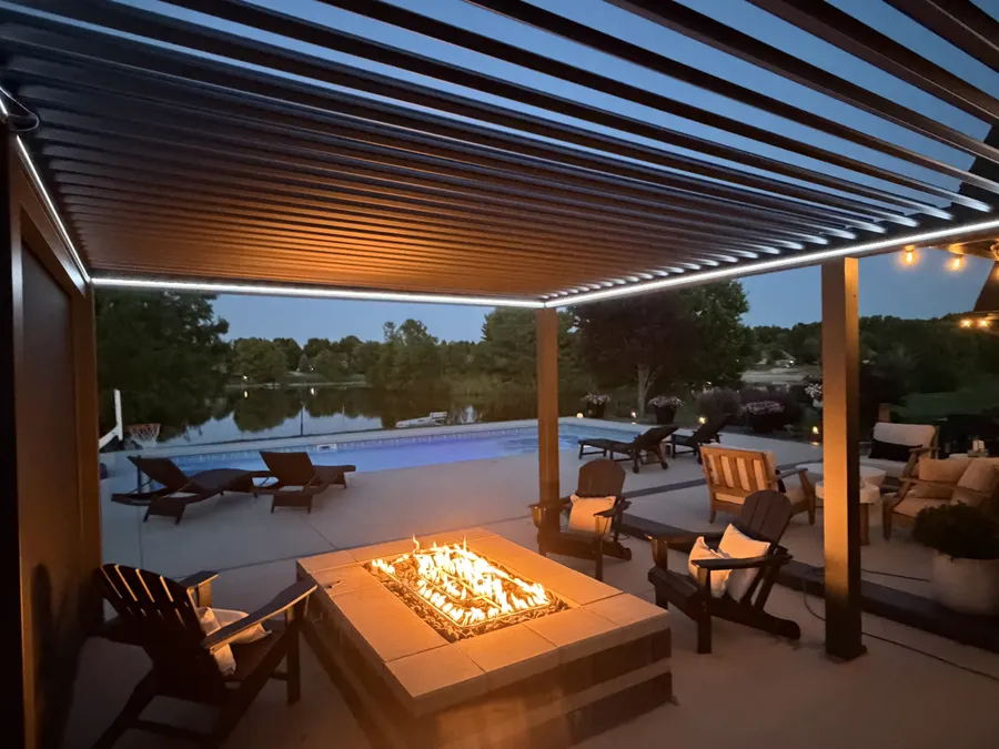 A finished louvered pergola with a fire pit and seating beside a pool, photographed in the evening.